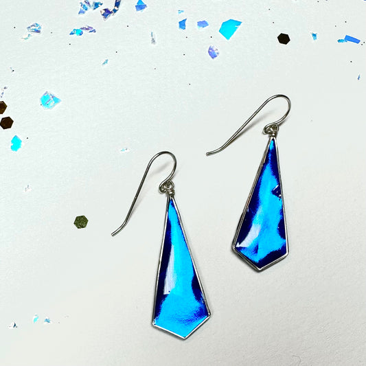 These bright metallic blue earrings are dramatically lit showing off their range of blues. Iridescent glitter surrounds the earrings to the top and left. Jay Bird Studio's jewelry is completely original and made in small batches.