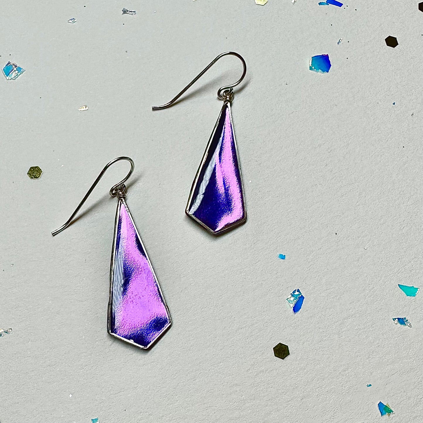 Metallic purple earrings in an elongated diamond shape that is created by hand in sterling silver square wire. Cream and a splash of party glitter as background because at JBS we celebrate you through bright, bold, one of a kind jewelry.