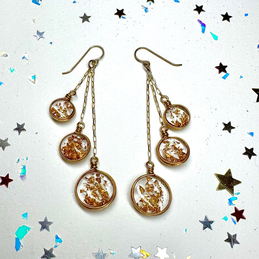 These earrings are party ready! Surrounded by silver and gold star glitter as well as iridescent party glitter. The dangle earrings feature transparent resin with three dimensional falling gold foil. Hanging off of fancy Sterling chain the baubles are three to an earring and taper from large on bottom to small on top.